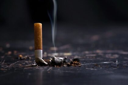 A cigarette butt with smoke rising from it, lying on a dark surface scattered with ashes.