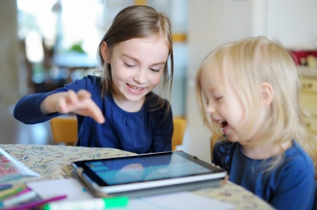 Two young children using a tablet while sitting at a table, both smiling and engaged.