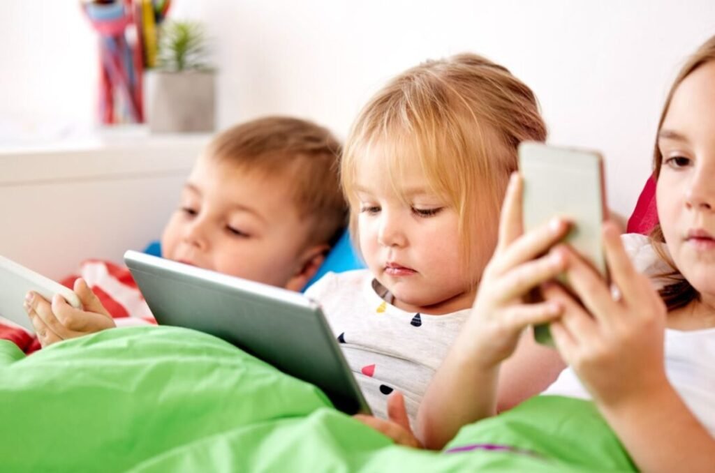 Three children lying in bed, focused on using electronic tablets and a smartphone.