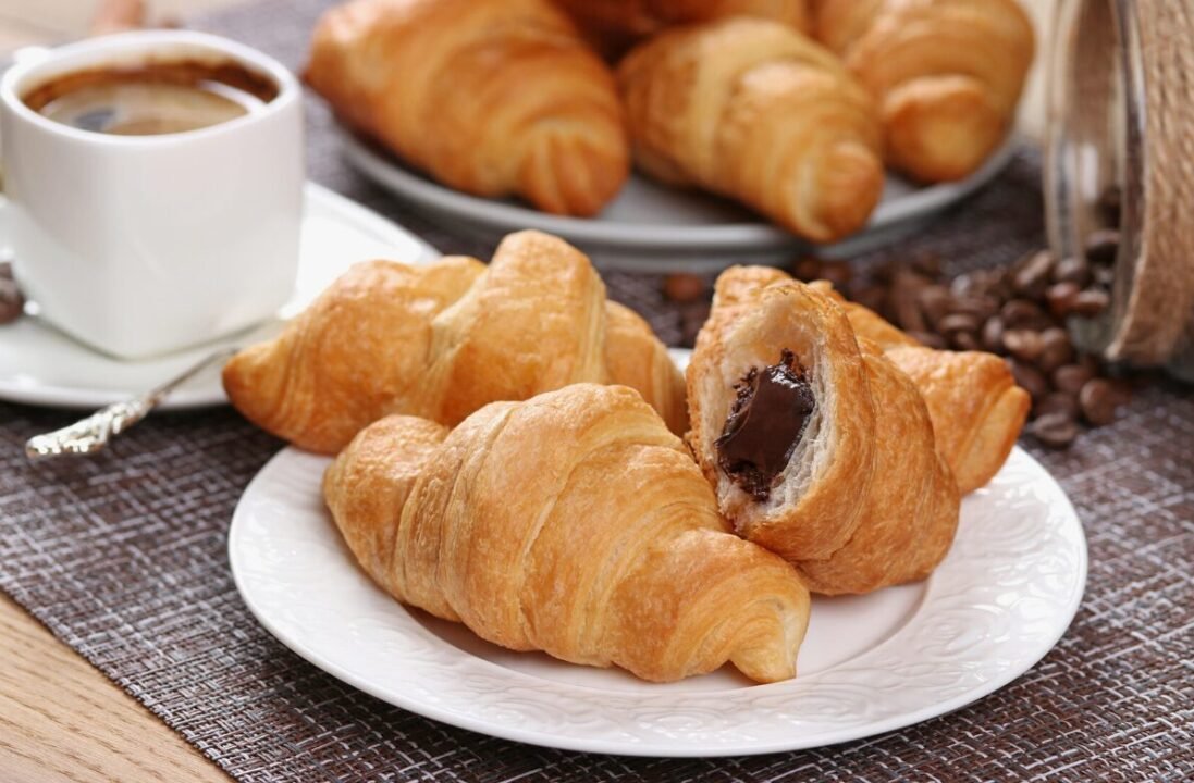 Freshly baked croissants with one split open revealing a chocolate filling, served alongside a cup of coffee and scattered coffee beans on a woven table mat.