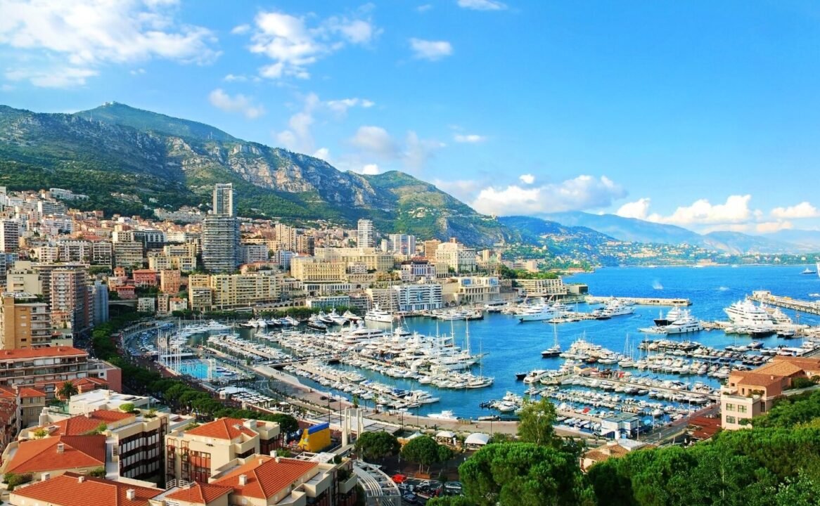 Luxurious coastal city with vibrant marina, mountains, and Mediterranean architecture under clear blue sky.