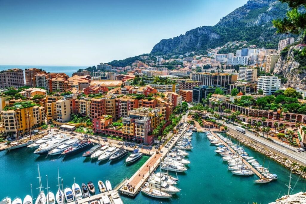 Aerial view of a vibrant marina surrounded by colorful buildings, with yachts docked in the water, alongside palm tree-lined streets and a mountainous backdrop.