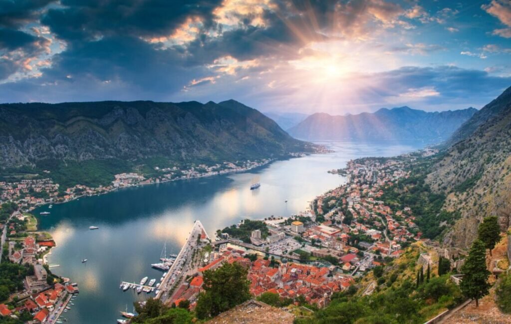 Aerial view of a coastal town nestled between lush mountains along a bay, featuring sun rays breaking through clouds over the landscape.