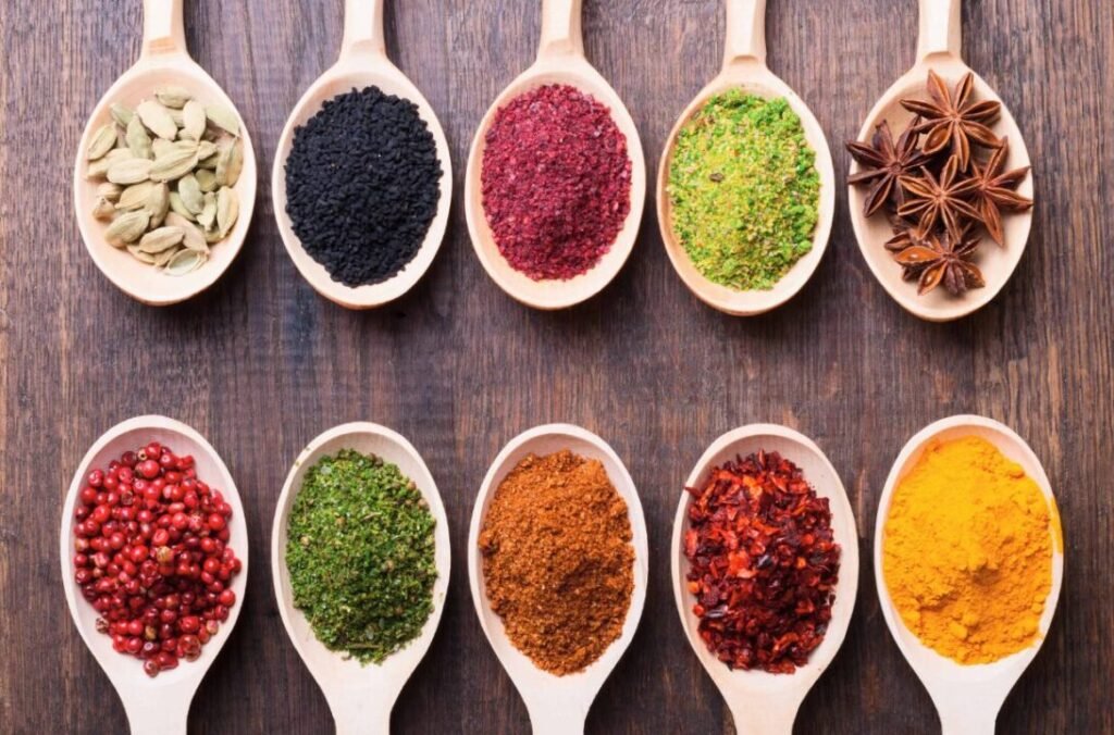 Variety of colorful spices and herbs in wooden spoons arranged on a wooden surface.