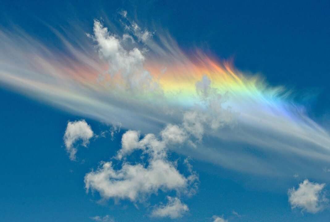Vibrant rainbow colors shine through cirrus clouds against a clear blue sky.