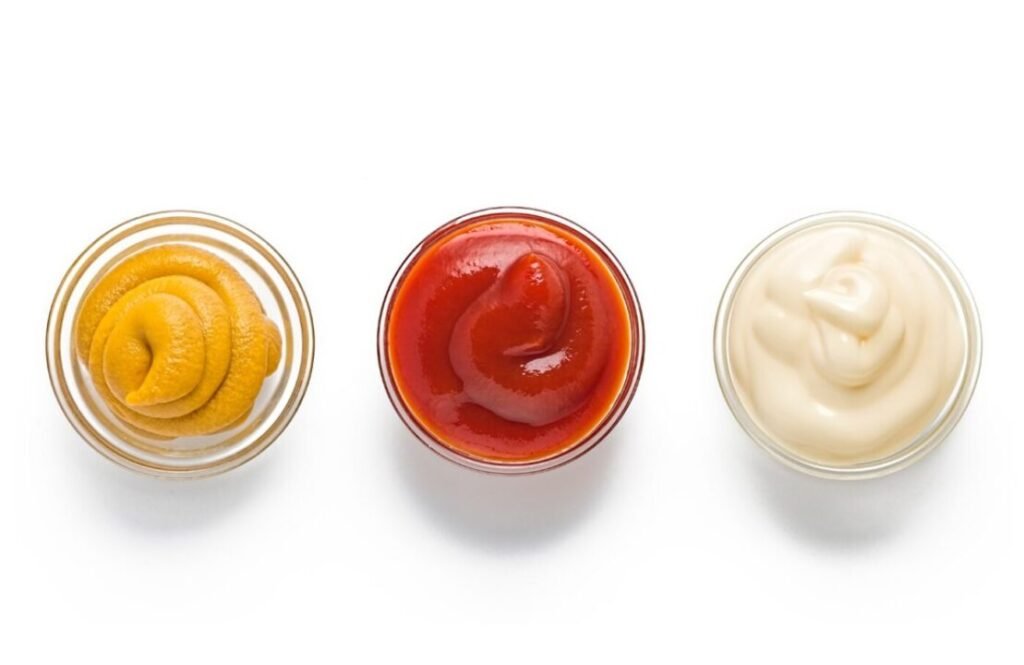 Three bowls of condiments isolated on white background, from left to right: mustard, ketchup, and mayonnaise.