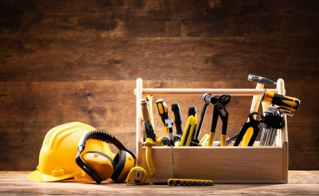 A variety of tools and a yellow hard hat with protective ear muffs on a wooden bench, all against a dark wood backdrop.