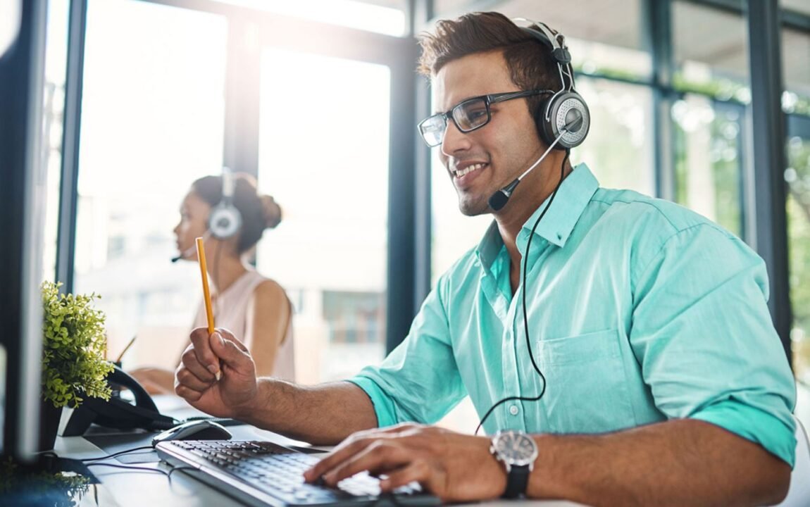 Man with glasses and headset smiling while working on computer in bright office setting.