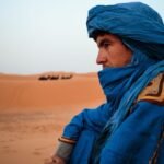 Tuareg person in blue robe gazes at sunset with resting camels in desert.