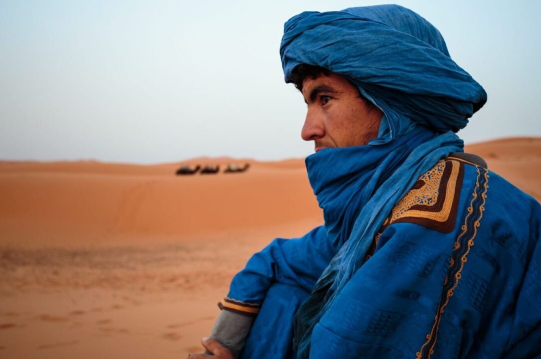 Tuareg person in blue robe gazes at sunset with resting camels in desert.