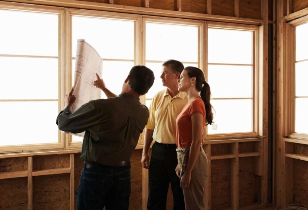 Three people, one holding a blueprint, discussing a project inside a framed, unfinished wooden house near large windows.