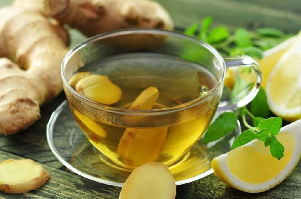 A clear glass cup of ginger tea with sliced ginger and lemon pieces on a wooden surface, surrounded by fresh ginger roots, lemon slices, and green leaves.