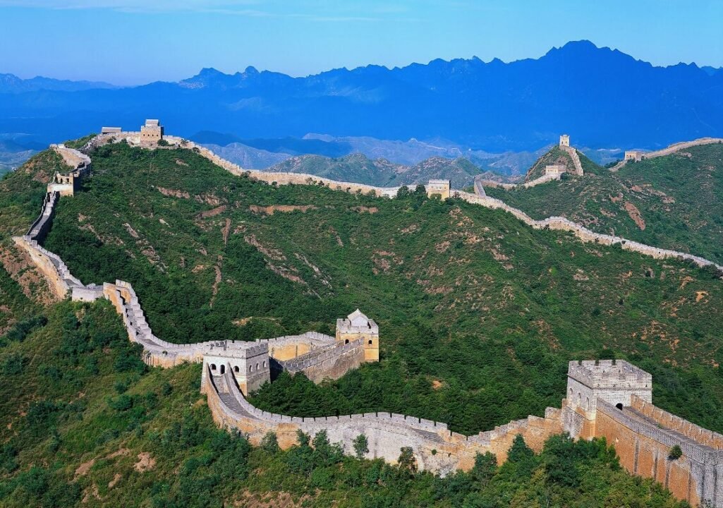 The Great Wall of China winds over green, mountainous terrain under a clear blue sky.