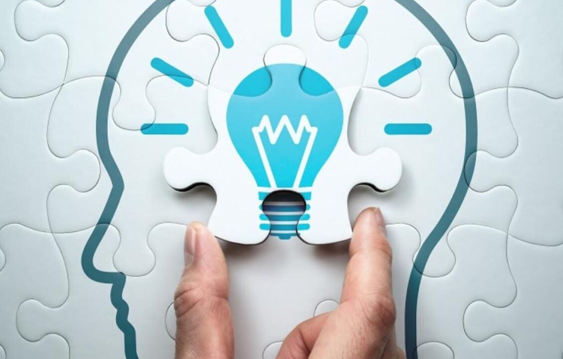 A hand placing a puzzle piece with an illuminated light bulb icon into a puzzle forming a human head outline.