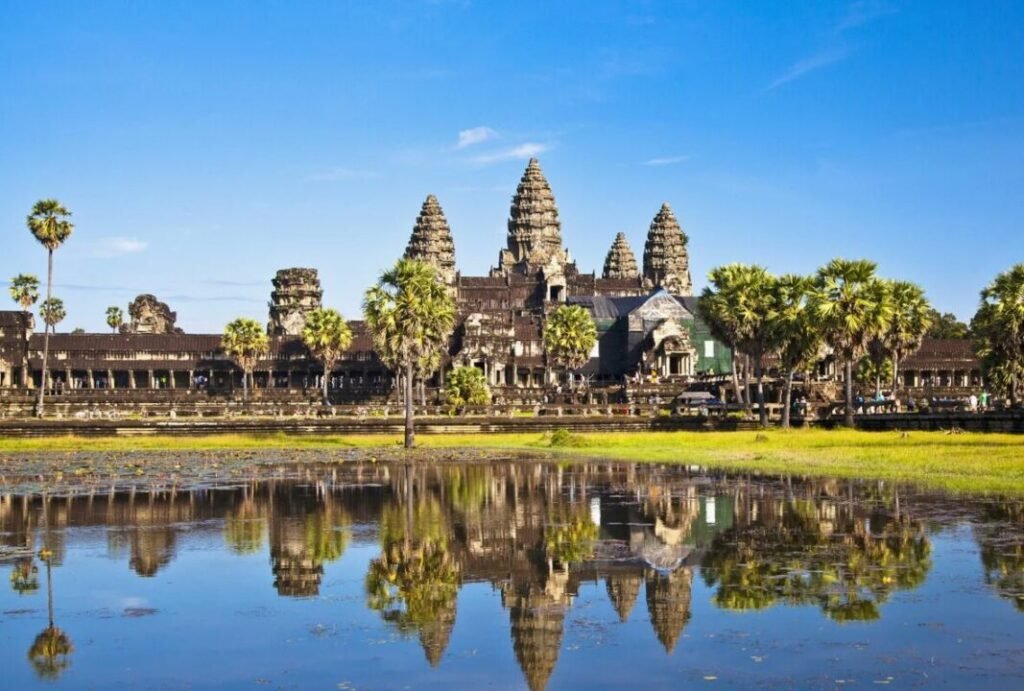 Angkor Wat temple complex in Cambodia, featuring the iconic towers and reflected in a large water pond, with clear blue sky and surrounding greenery.