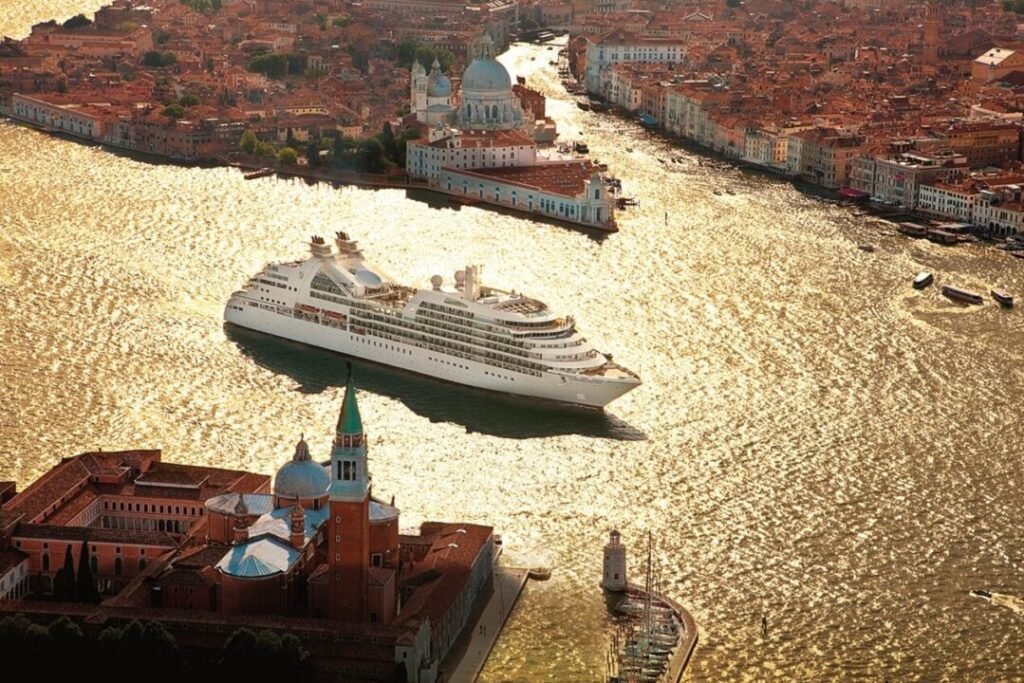 Aerial view of a large cruise ship in the glittering waters near Venice, with historic buildings and the cityscape in the background.