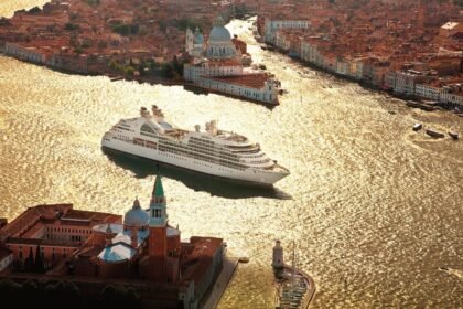 Aerial view of a large cruise ship in the glittering waters near Venice, with historic buildings and the cityscape in the background.