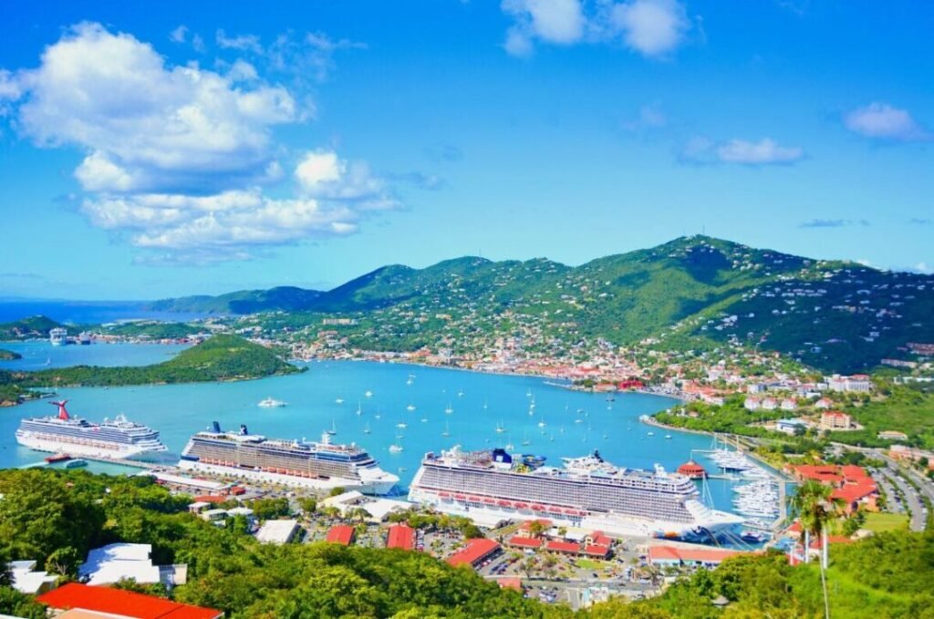 Aerial view of a vibrant harbor with multiple cruise ships docked, surrounded by lush green hills and a clear blue sky.