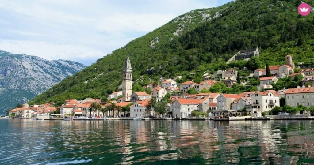 Scenic view of a coastal town with historic buildings and a towering church spire, nestled between lush green mountains and a calm sea.