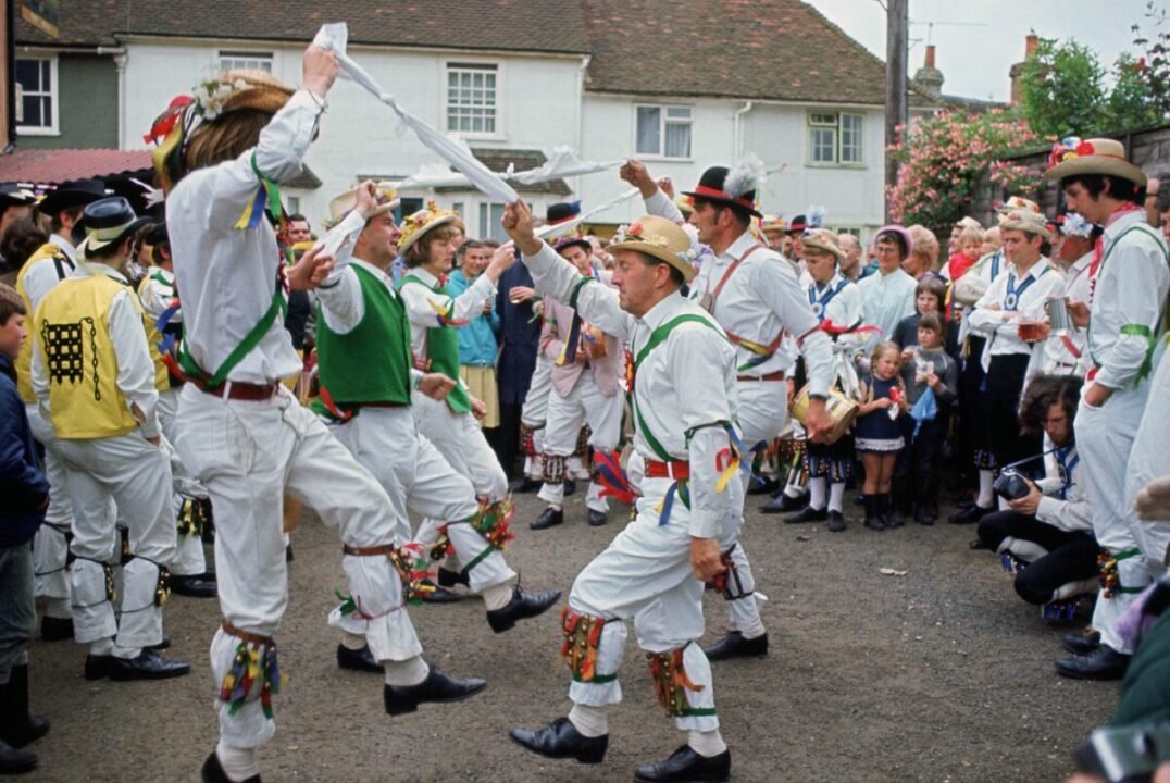 Men dressed in traditional Morris dancing attire perform a dance with white handkerchiefs, while a crowd watches in a village setting.