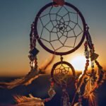 Dreamcatcher with feathers and beads held against a sunset sky.