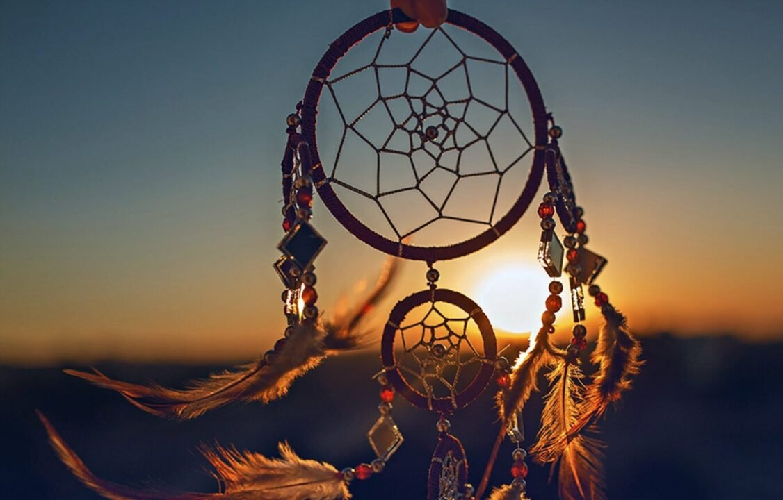 Dreamcatcher with feathers and beads held against a sunset sky.