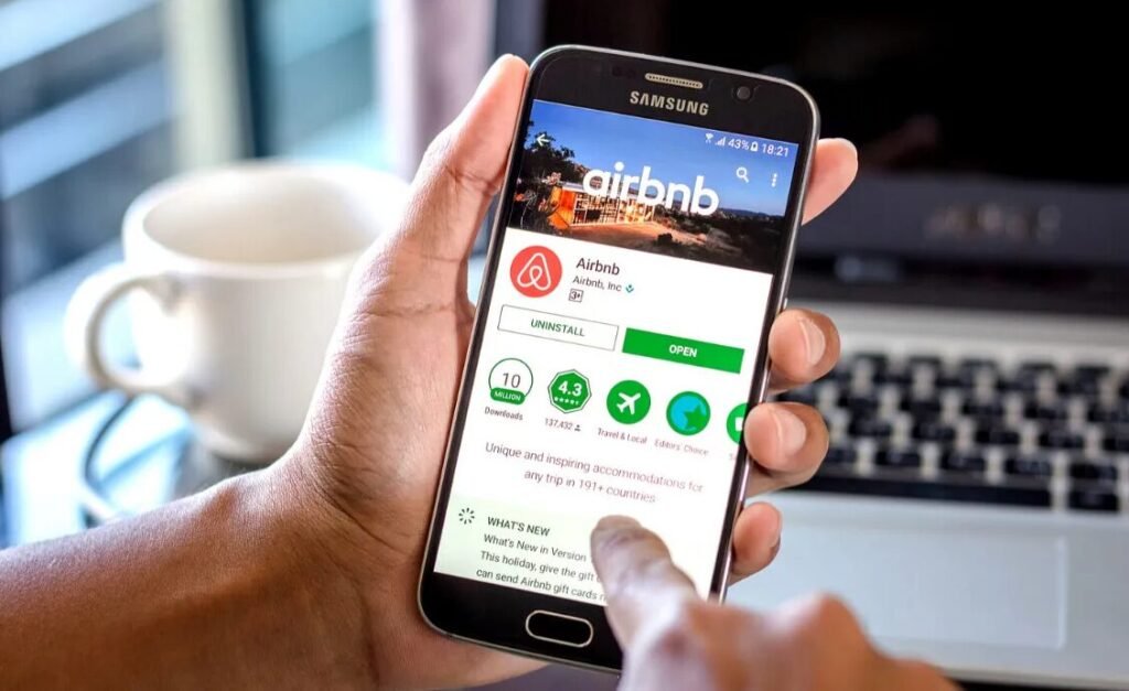 Person using an Airbnb app on a Samsung smartphone with a laptop and coffee cup in the background.