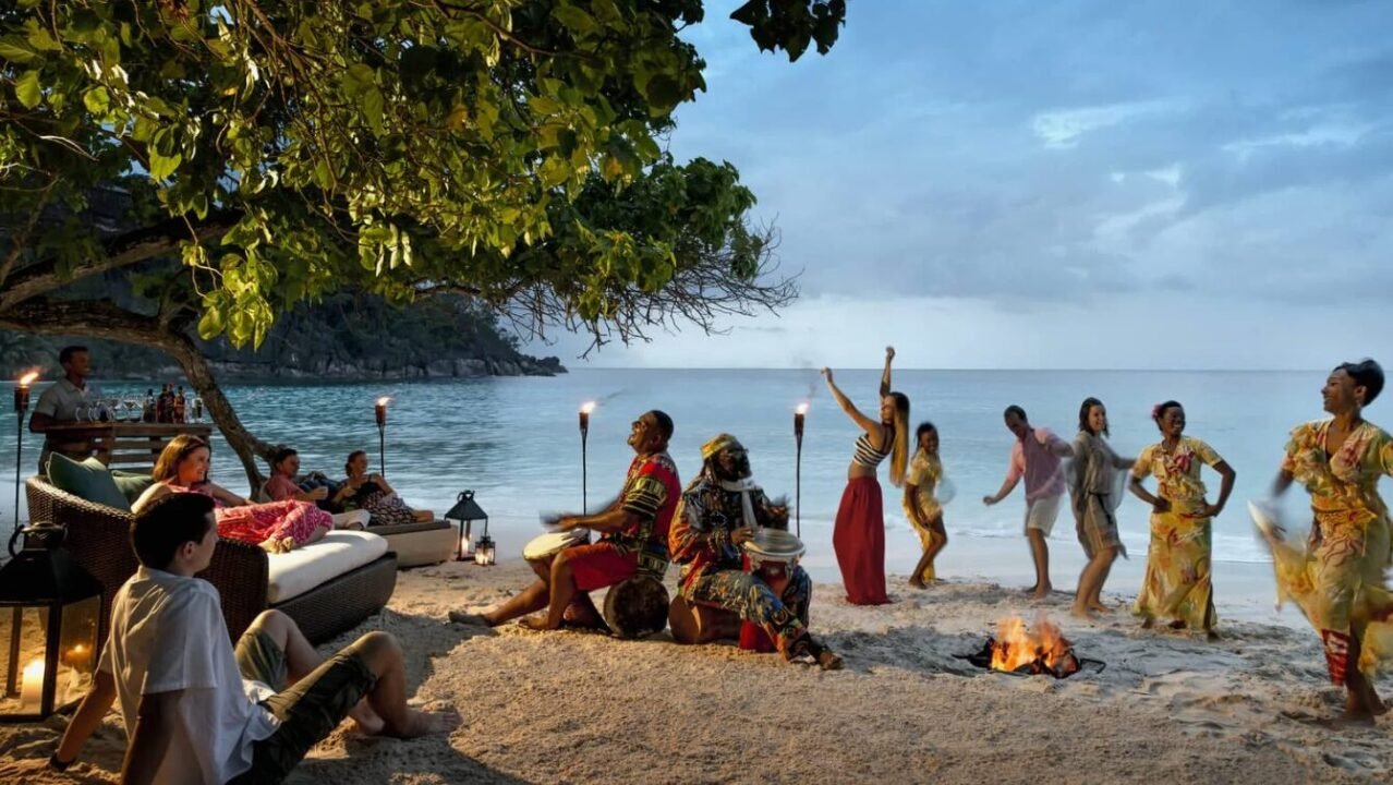 People relaxing and watching dancers around a bonfire on a beach at dusk, with musicians playing drums under a tree.