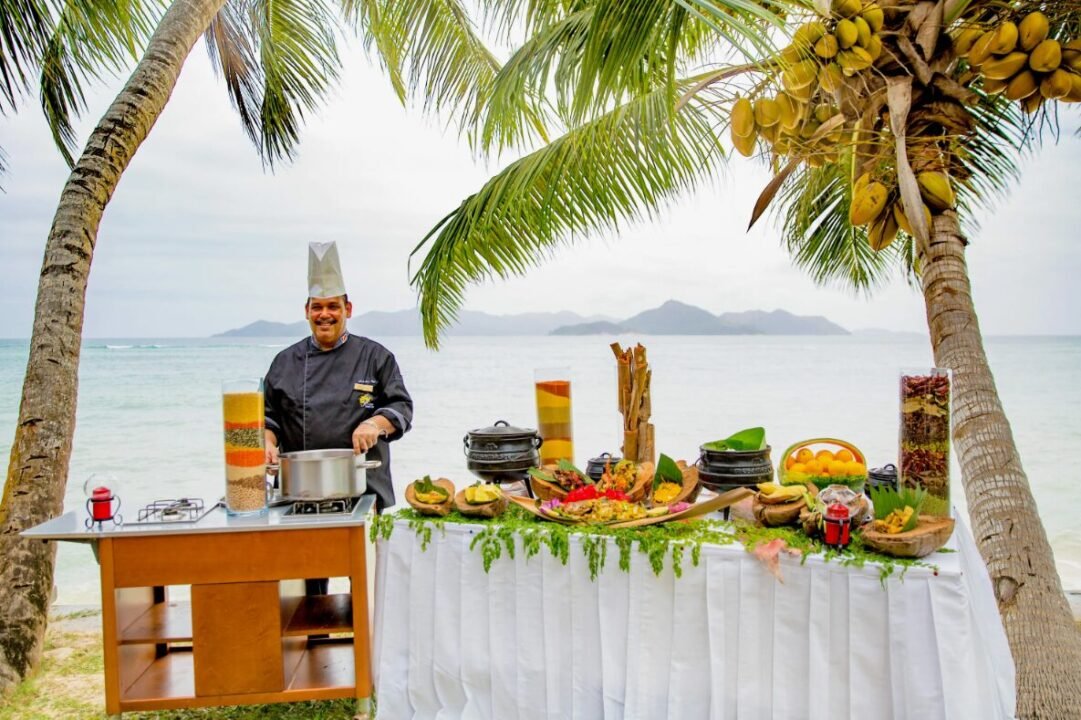 Chef in uniform and hat cooking at an outdoor setup by the beach, with a variety of food and ingredients on a table, surrounded by palm trees.