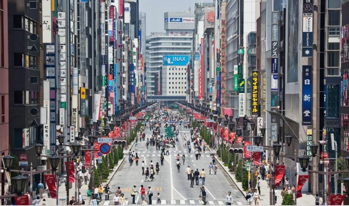 Crowded urban street in Tokyo with tall buildings on either side, covered in colorful advertisements and signs in Japanese.