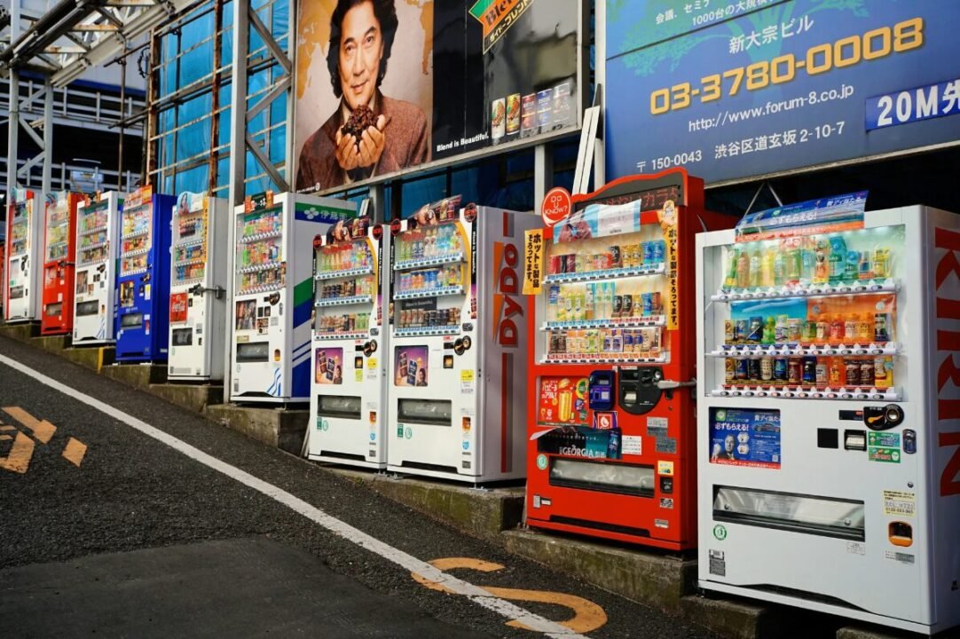 A row of colorful vending machines on a slightly inclined sidewalk with advertisements on the wall behind them.