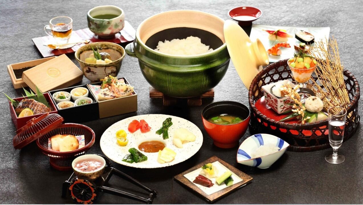 A traditional Japanese meal laid out on a black surface, featuring various dishes including sushi, rice in a green pot, miso soup, tempura, pickles, assorted side dishes, and beverages in cups and glasses.