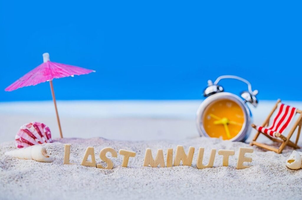 Miniature beach scene with the phrase "LAST MINUTE" in wooden letters, a pink parasol, a seashell, a striped deck chair, and an alarm clock on a sandy surface against a blue background.