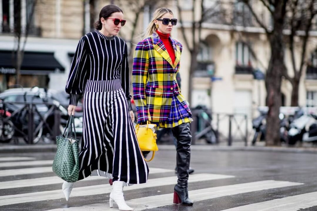 Two stylish women walking on a city street, one wearing a black and white striped dress and the other wearing a yellow plaid jacket and over-the-knee boots.