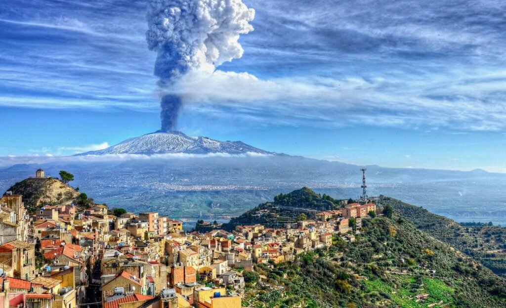 A scenic view of a town with colorful houses in the foreground, lush green hills, and a smoking Mount Etna volcano in the background under a bright blue sky.