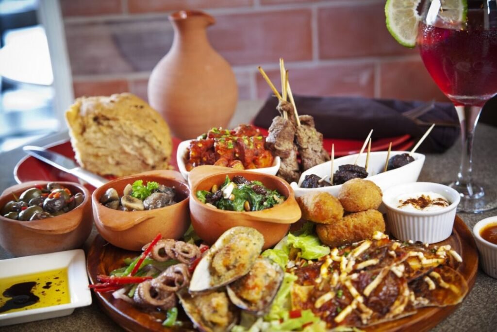 A variety of tapas dishes including olives, mushrooms, salad, bread, skewered meat, croquettes, and mussels, served on a wooden platter with dips and a drink.