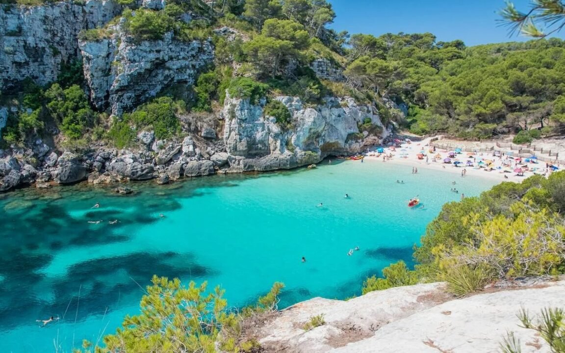 Aerial view of a picturesque beach with clear turquoise water, surrounded by rocky cliffs and lush green trees. The beach is dotted with colorful umbrellas and people sunbathing and swimming.