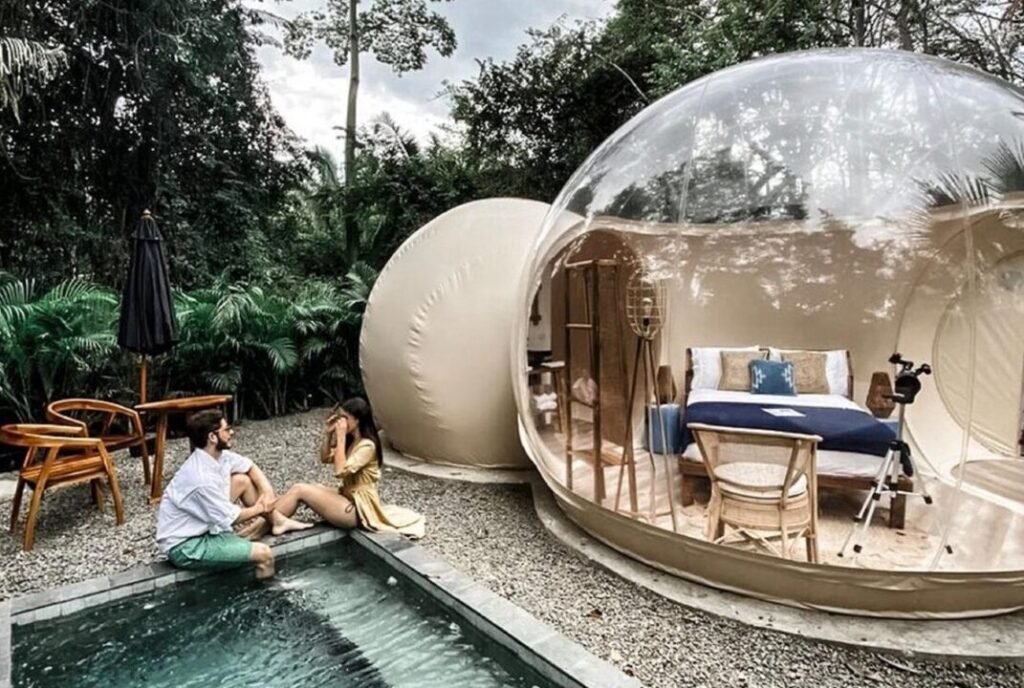 A couple sitting by a small pool outside a transparent bubble tent furnished with a bed and chairs, surrounded by dense green foliage.