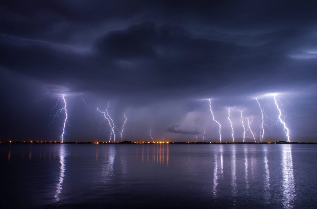 Dramatic night sky with multiple lightning bolts over calm water and distant shore lights.