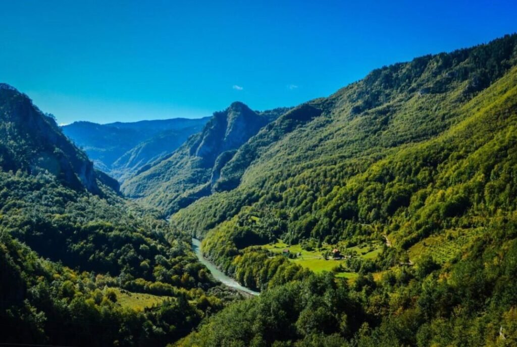 Scenic view of a lush green valley with a winding river, surrounded by steep forested hills under a blue sky.