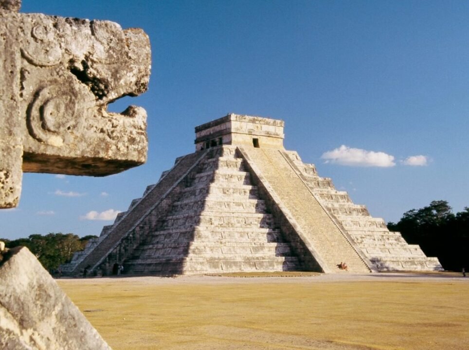 El Castillo pyramid at Chichen Itza with a close-up of a carved stone in the foreground, under a clear blue sky.