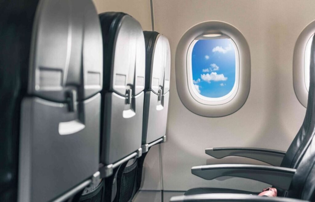 Interior view of an airplane showing empty seats, overhead compartments, and a clear view of blue sky with clouds through the window.