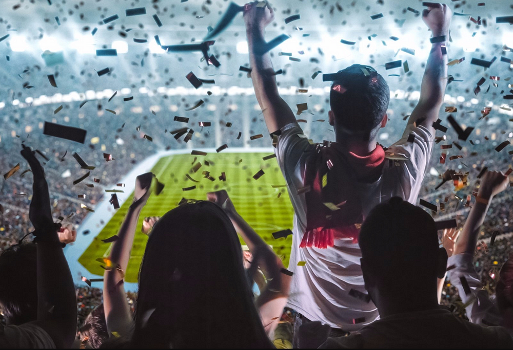 Crowd of spectators with raised arms celebrating at a stadium with confetti falling during a nighttime event.