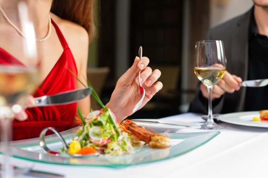 Two people dining at a restaurant with elegant table settings, eating gourmet meals and drinking white wine.