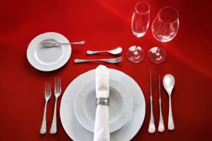 Formal table setting with white plates, a napkin ring, cutlery, and two wine glasses on a red tablecloth.