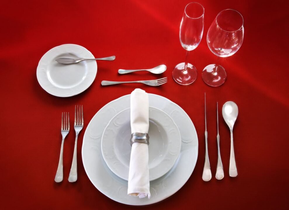 Formal table setting with white plates, a napkin ring, cutlery, and two wine glasses on a red tablecloth.