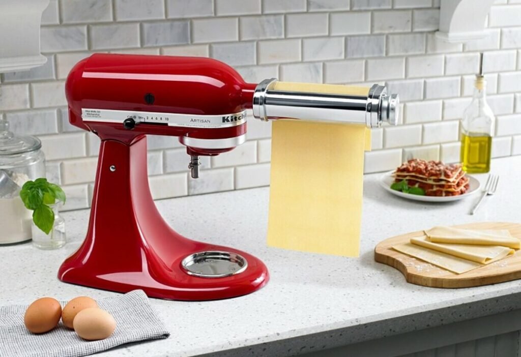 Red KitchenAid stand mixer with pasta maker attachment rolling out yellow dough on a kitchen counter, with eggs on a cloth and a meal of pasta in the background.