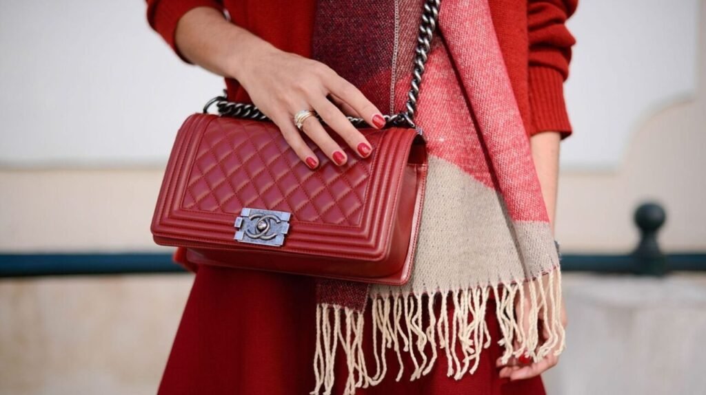 Close-up of a woman's hand holding a red quilted leather handbag with a silver logo clasp, complemented by a red outfit and a multicolored scarf with fringe.