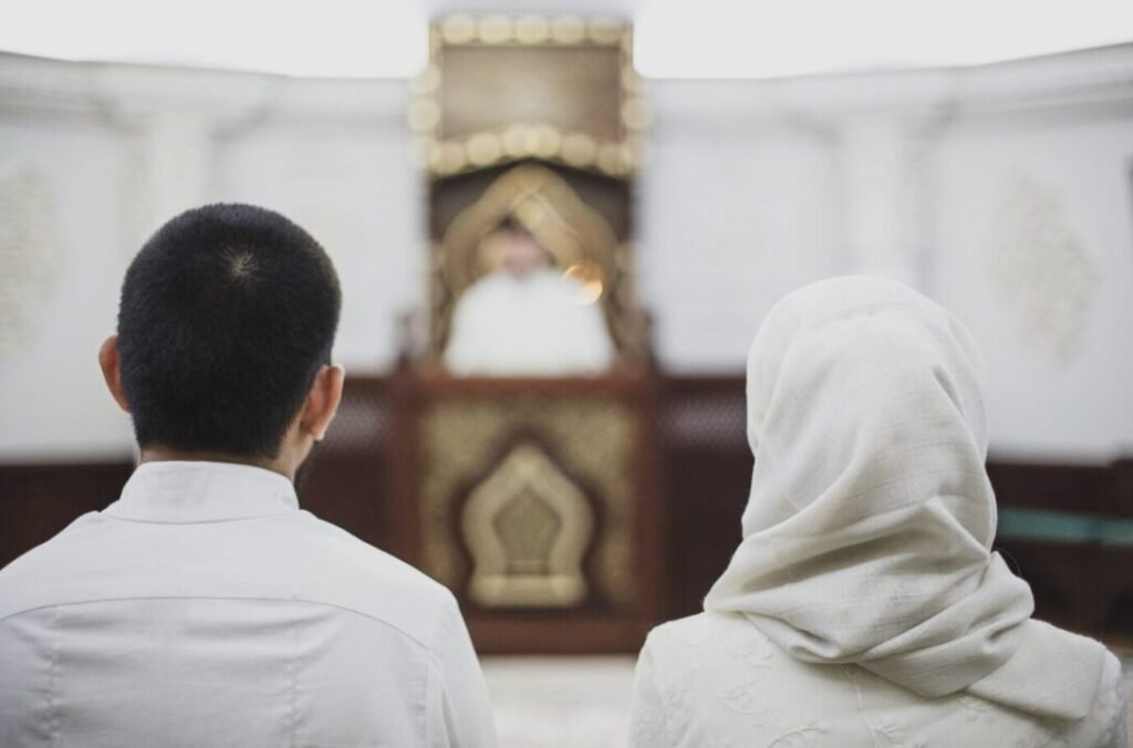 A man and a woman in white clothing, viewed from behind, sitting and facing a religious leader speaking at the pulpit in a mosque.