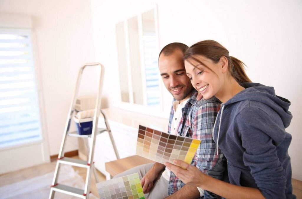 A couple examining paint color samples together in a room with painting supplies visible in the background.